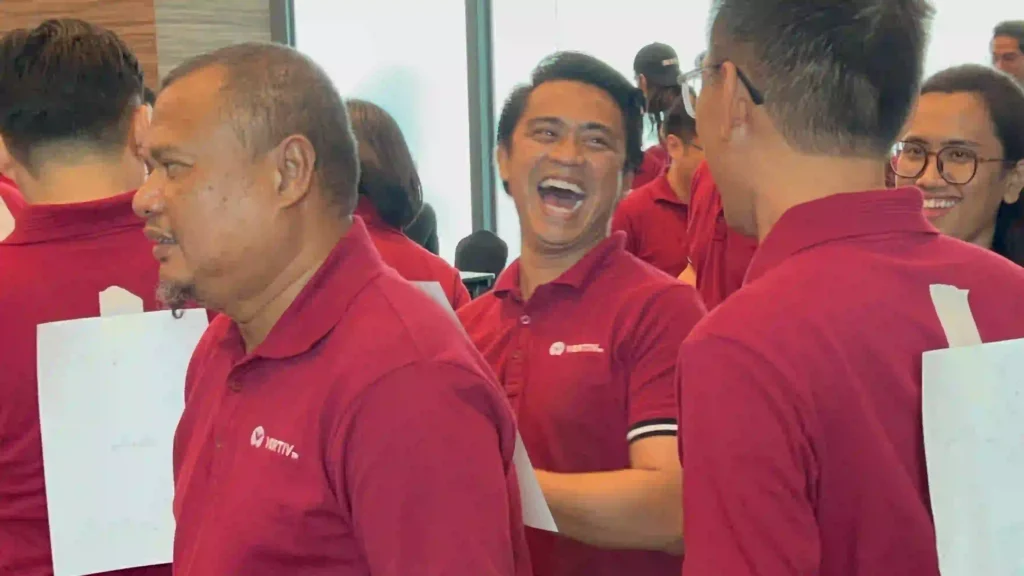 A group of customer in Vision Building event, wearing matching red polo shirts, are seen laughing and smiling during a team-building activity. One man in the center is captured mid-laugh, conveying a strong sense of pure fun.